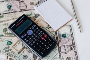 On a table, there are some banknotes, a calculator, a notepad, and a pen.