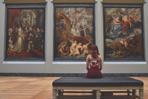 A girl sitting on a bench and looking at the paintings on the wall.